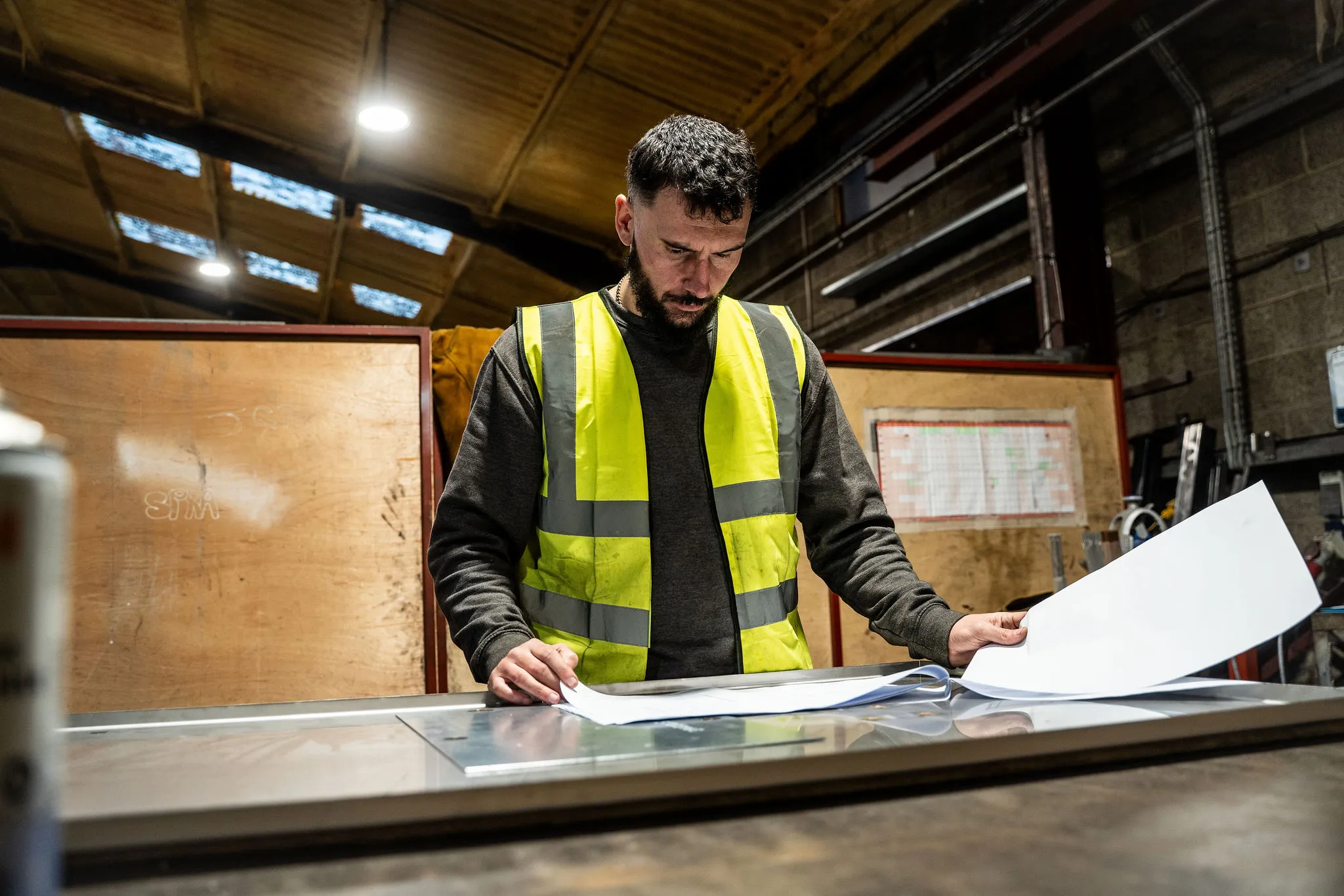 Man in high-visibility vest reviewing blueprints on a metal table in an industrial workshop.