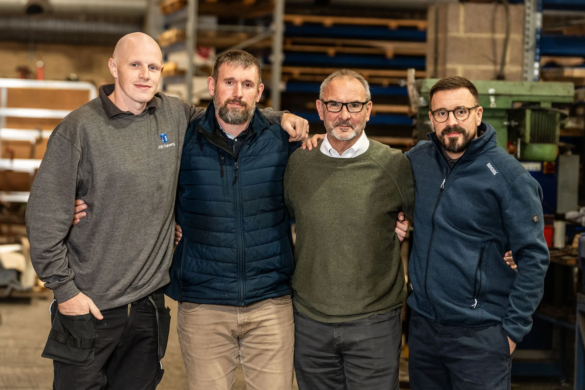 Four men standing arm in arm in a workshop, wearing casual and work jackets.