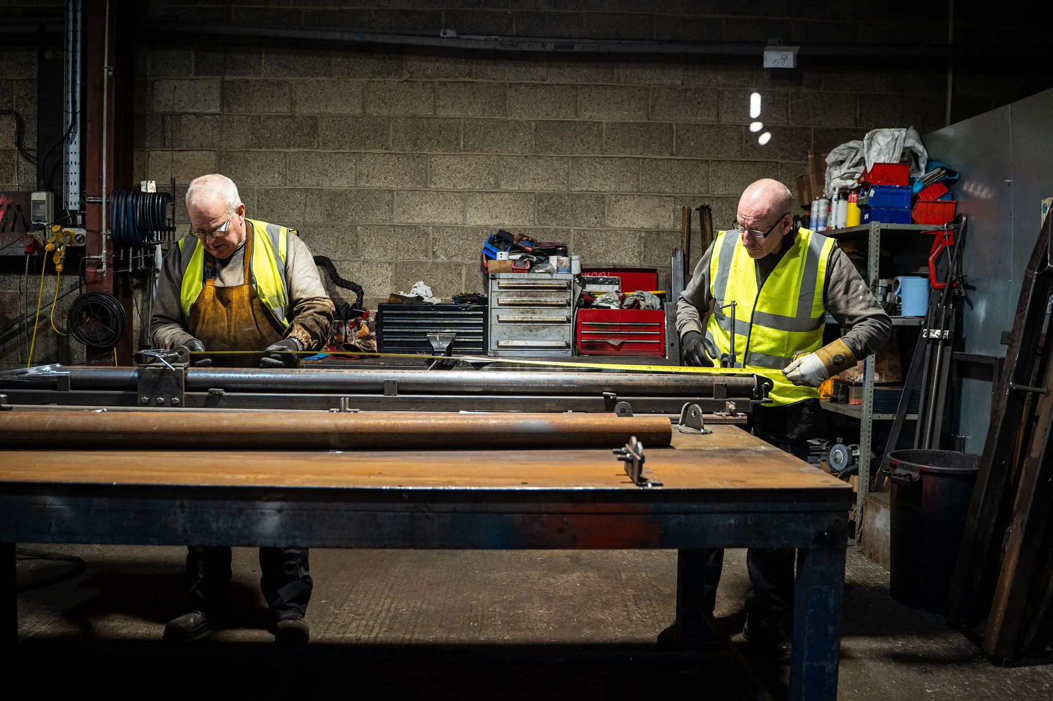 Two workers in yellow safety vests working with metal pipes at a workshop table.