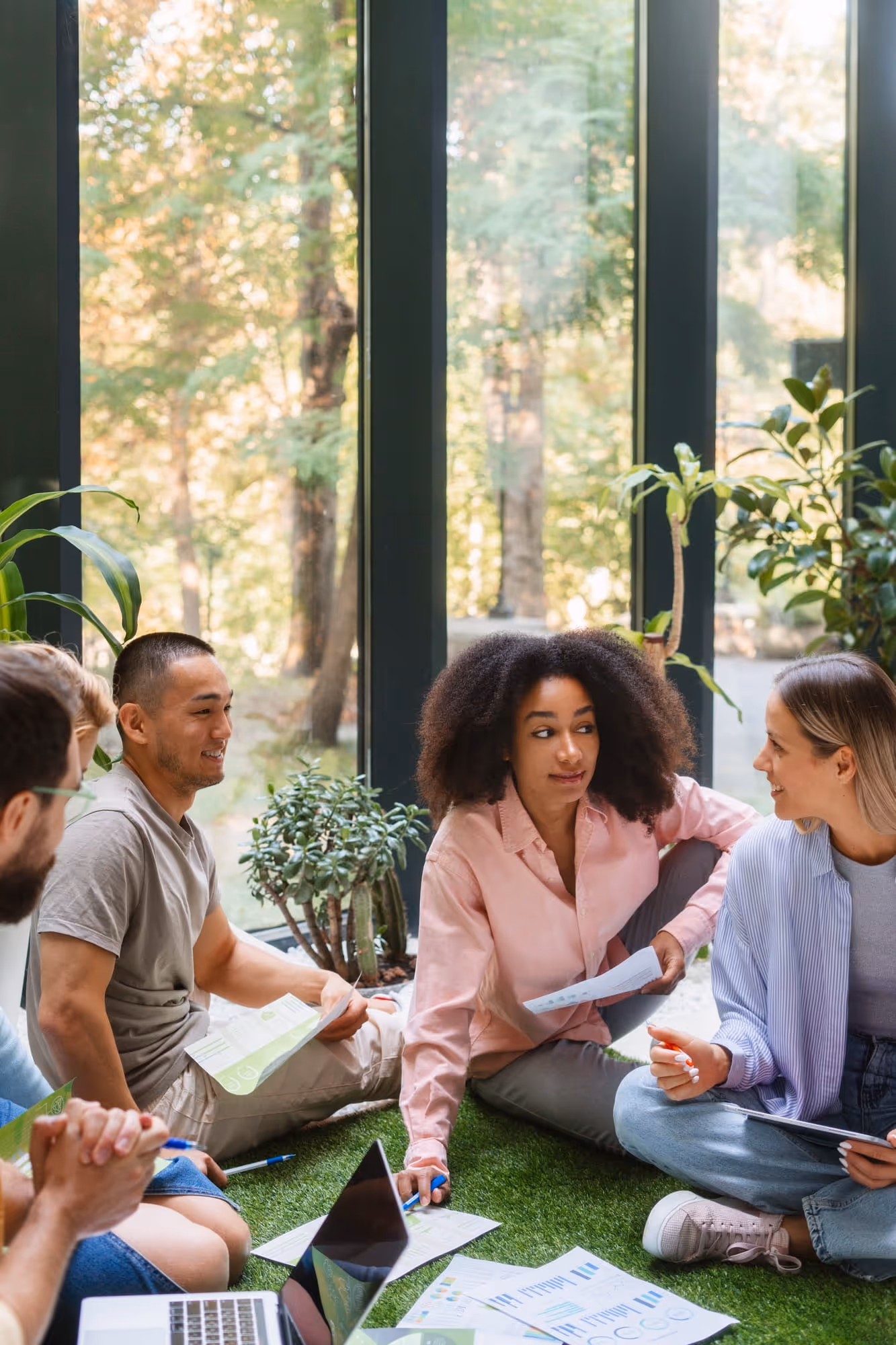 A diverse group of young adults sitting on grass indoors near large windows, discussing documents and working together.