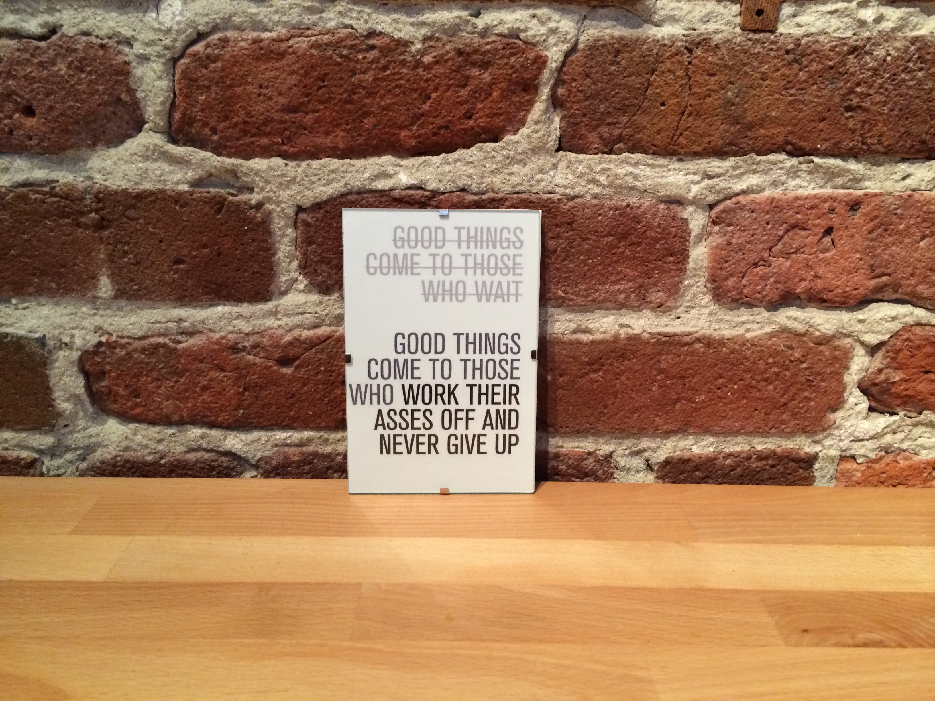 Sign on wooden table in front of a brick wall reading: 'Good things come to those who work their asses off and never give up.'