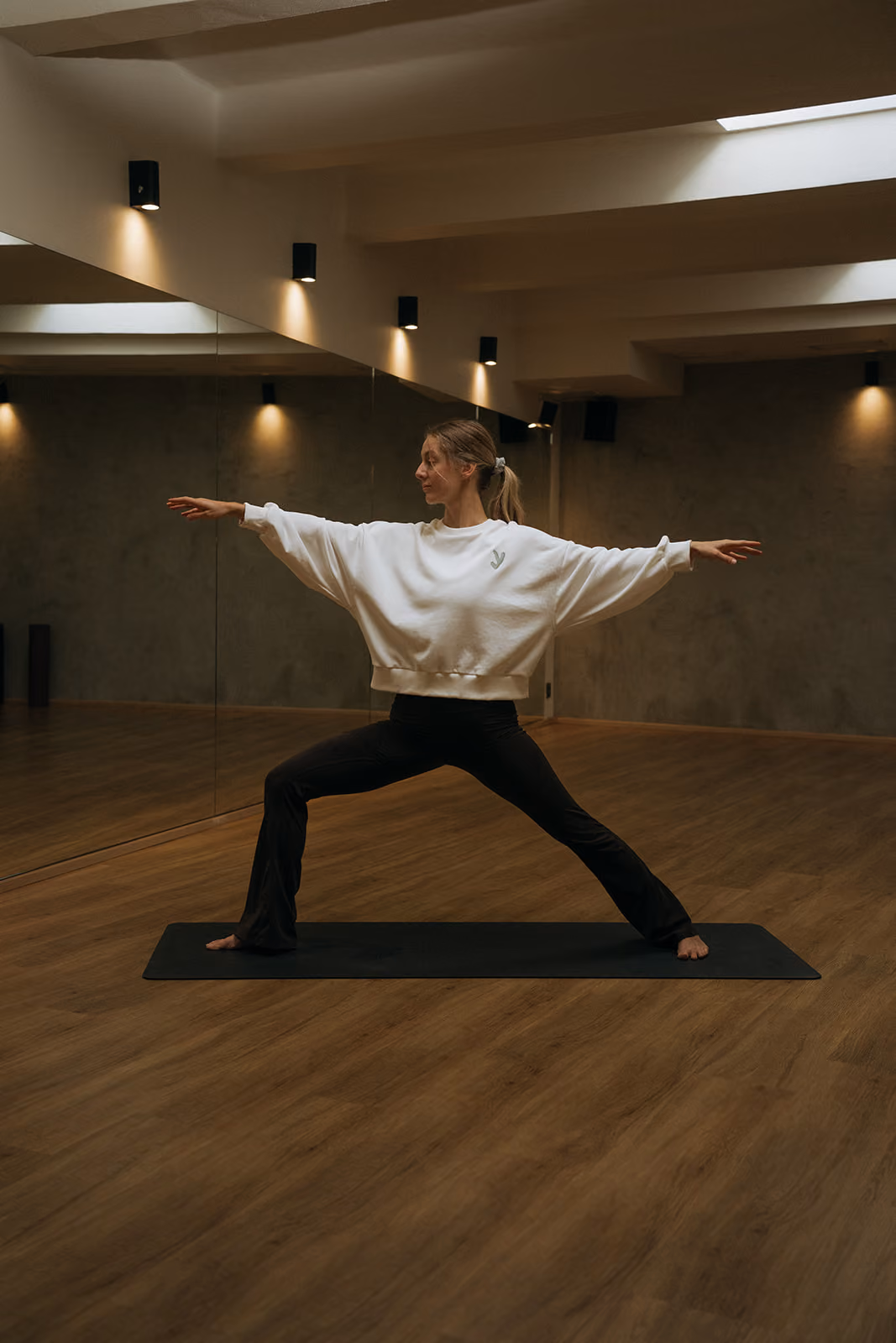 Celkový pohled na ženu v bílé jógové mikině v pozici bojovníka v interiéru studia Yoga Movement.