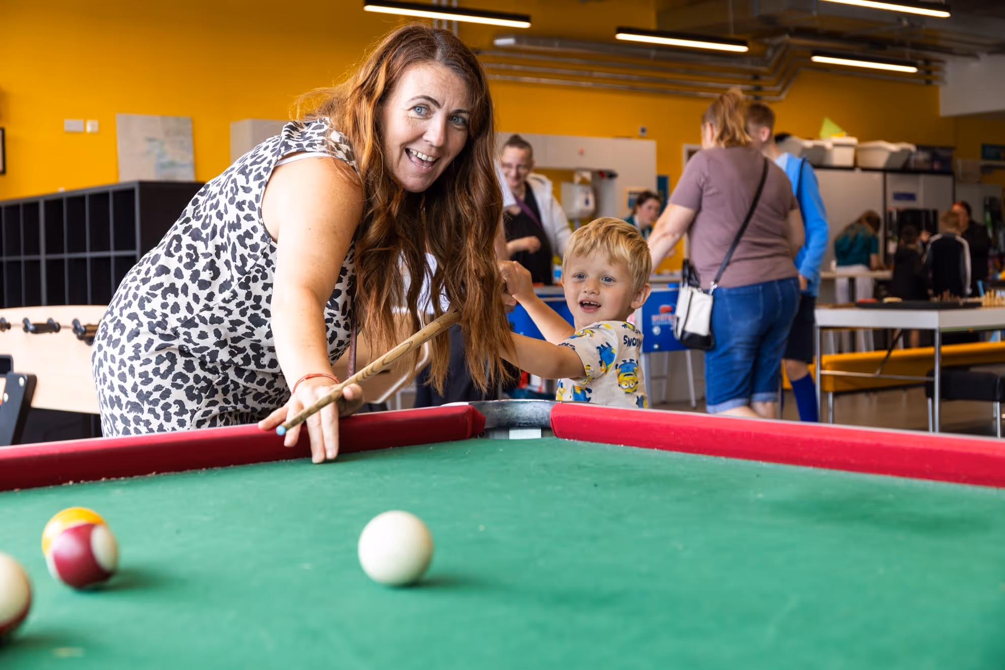 A woman with long hair smiling and playing pool with a young boy in a busy indoor recreational room.