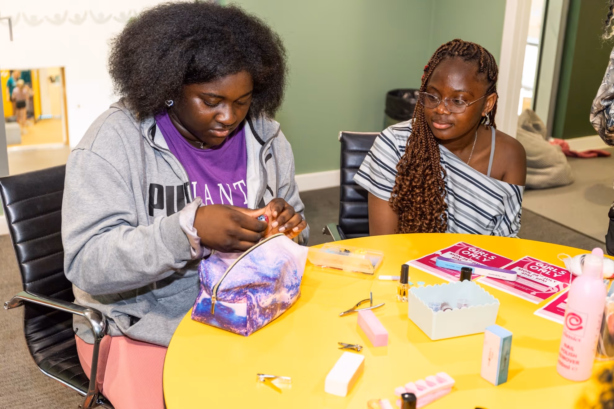 Two young women sitting at a yellow table with nail care tools, one woman holding a nail polish bottle while the other watches.