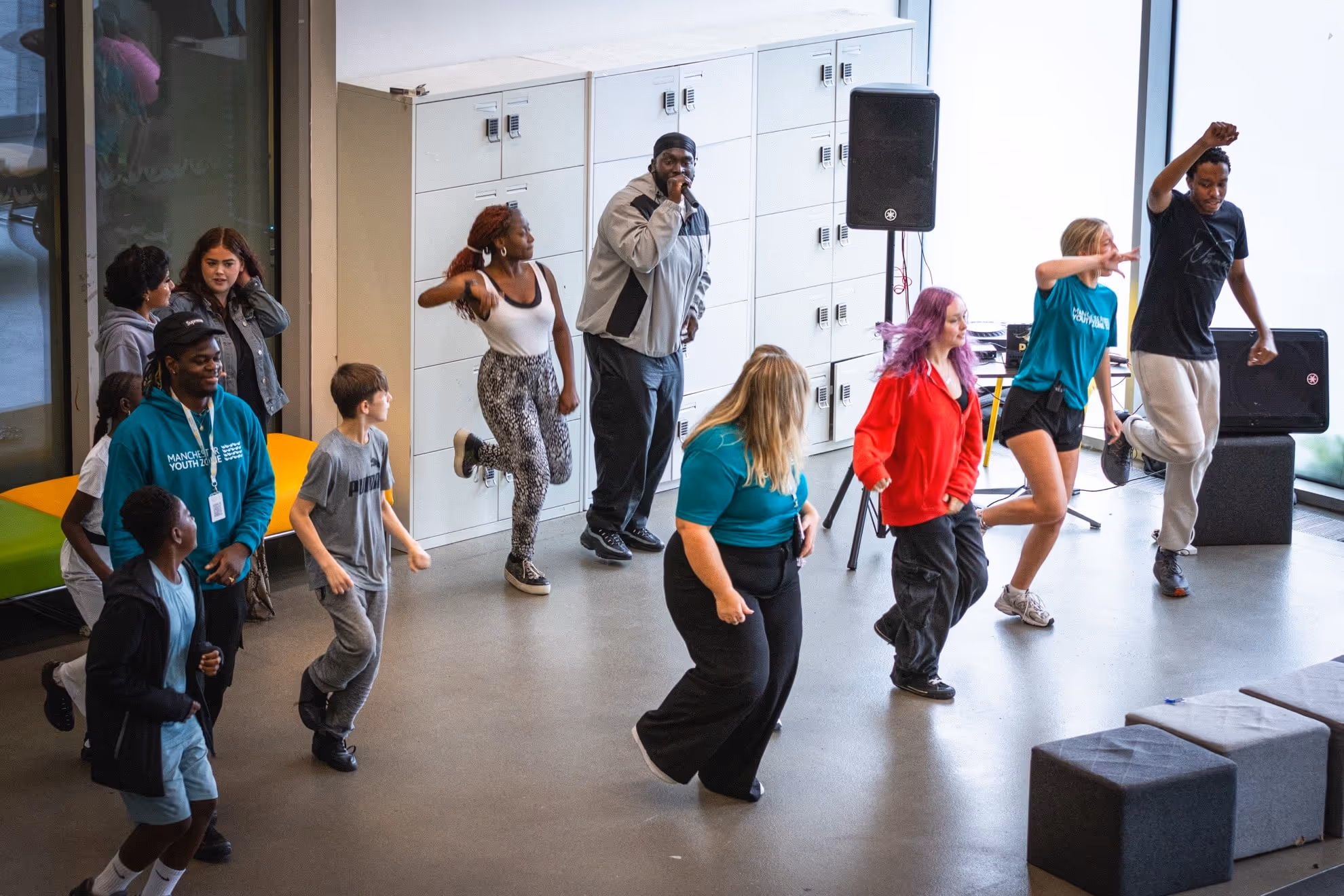 Group of diverse young people indoors dancing and singing near lockers and windows.