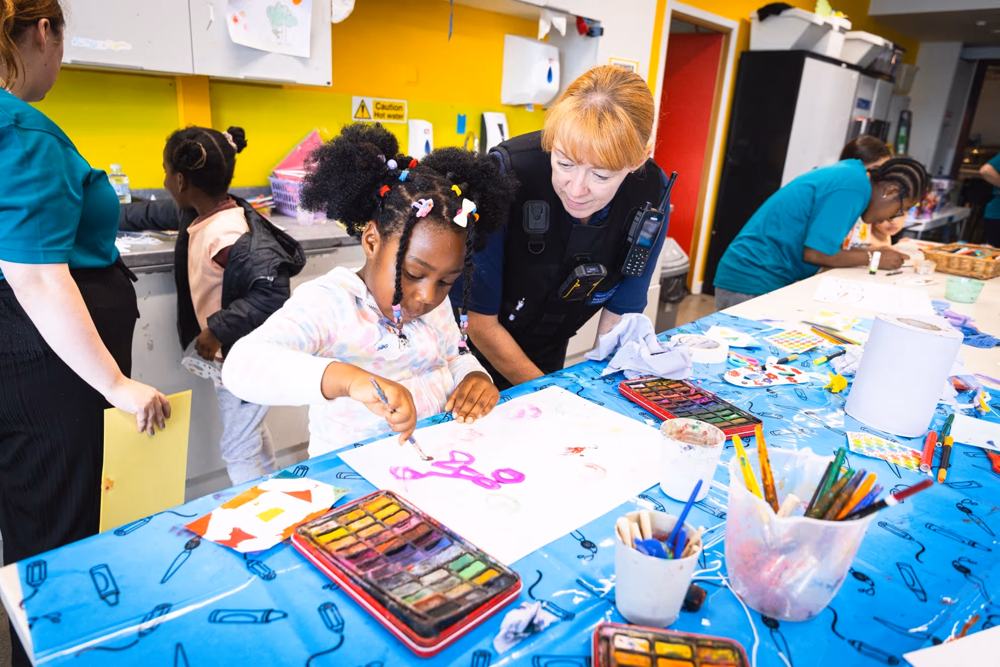 A young girl painting with purple watercolor while a police officer watches her at a craft table covered with art supplies.