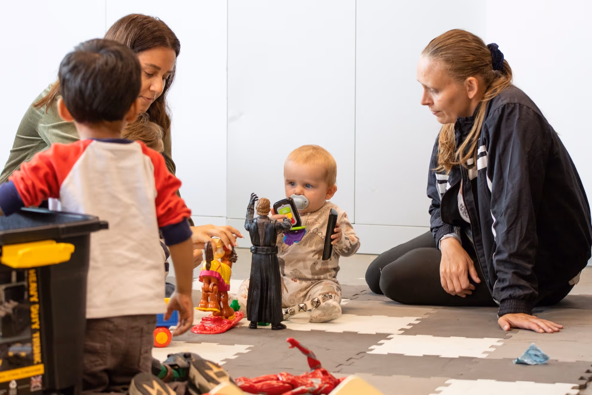 Two women and two children playing with toys on a padded floor mat in a bright room.