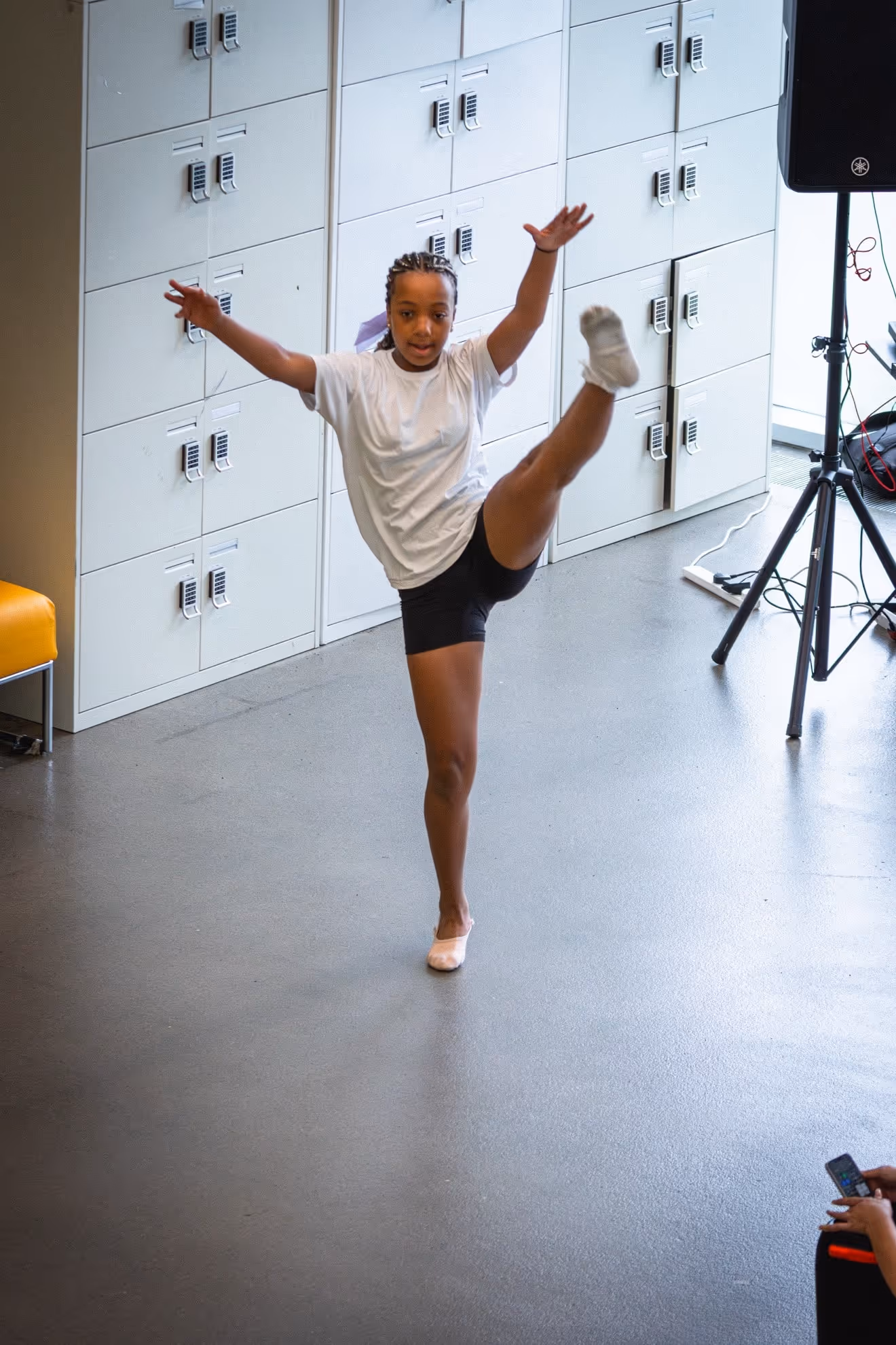 A young dancer in a white shirt and black shorts performing a high leg lift in a room with white lockers.