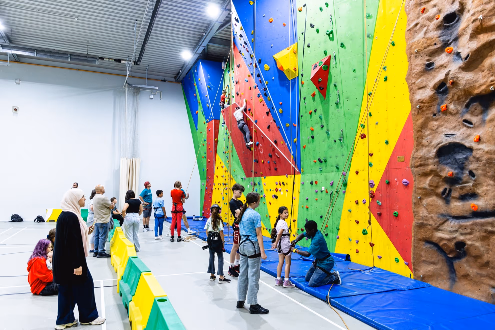 Indoor climbing gym with colorful climbing walls and children preparing or climbing while adults observe.