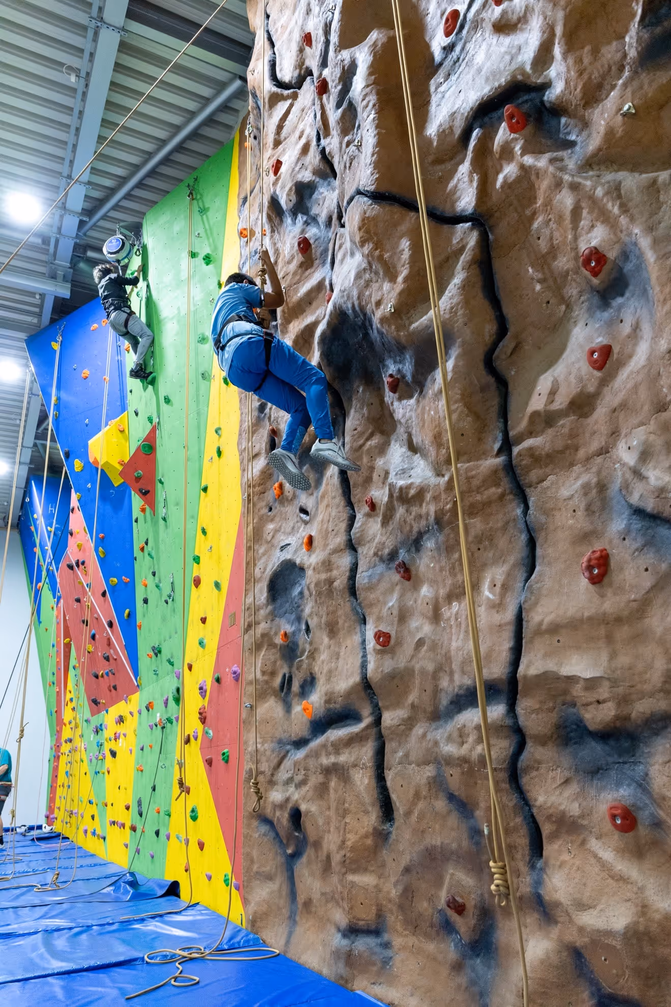 Two people climbing colorful and rock-patterned indoor climbing walls with safety ropes.