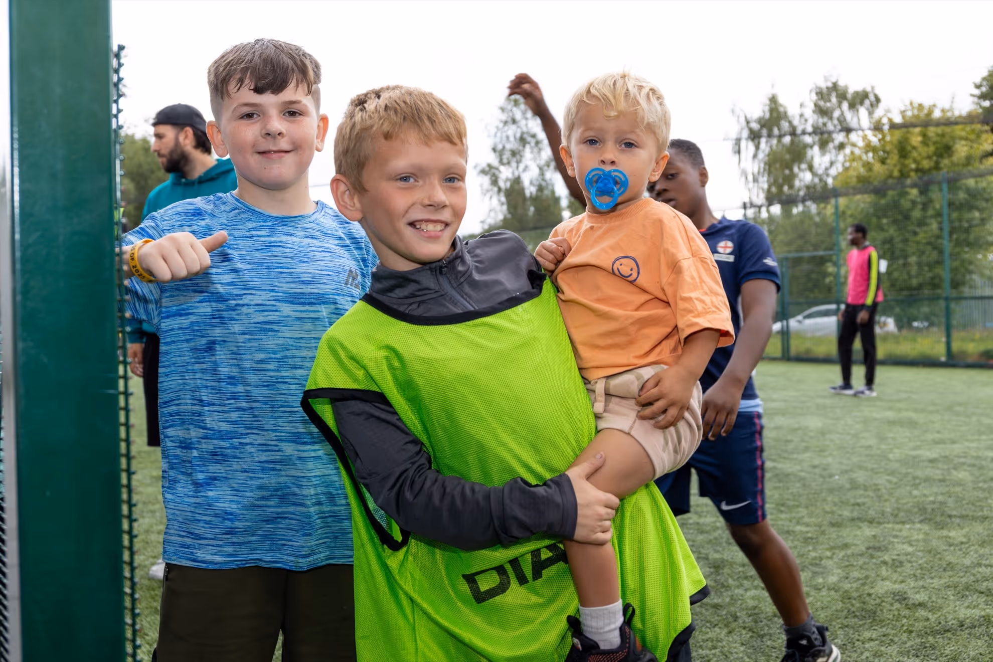 Two boys smiling at the camera, one holding a toddler with a pacifier, on a sports field with other people in the background.