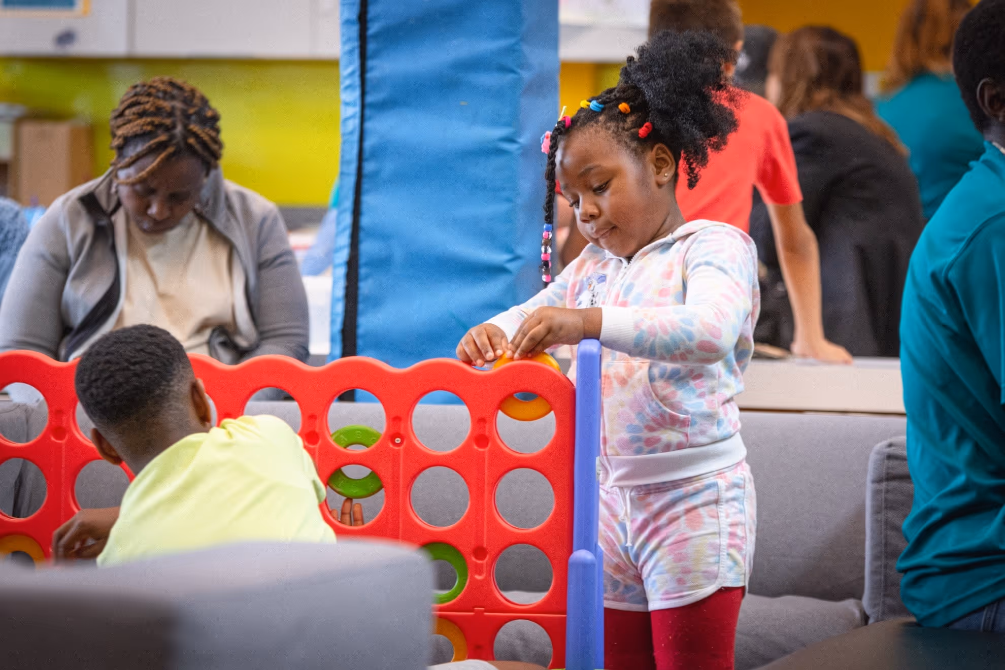 Two children playing a large Connect Four game indoors with adults seated and standing in the background.
