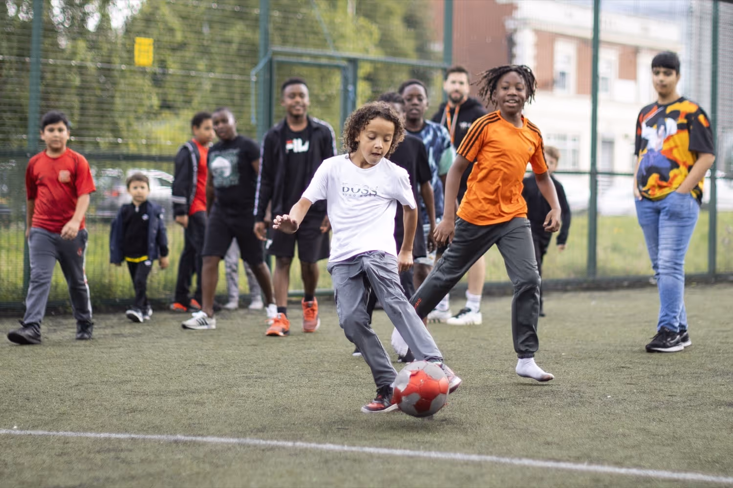 Children playing soccer on a grass field, with one child in a white shirt kicking a red and gray ball while others watch and run.