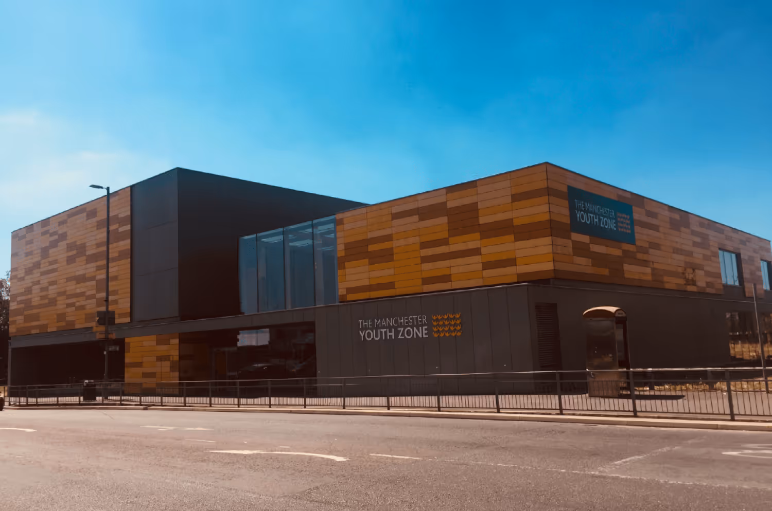 Modern building with wood paneling and dark lower facade labeled 'The Manchester Youth Zone' under a blue sky.