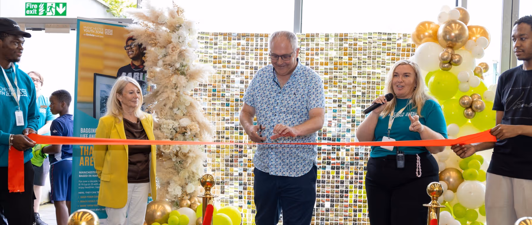 Group of people at a ribbon-cutting ceremony, with a man cutting a red ribbon and others holding it, surrounded by floral and balloon decorations.