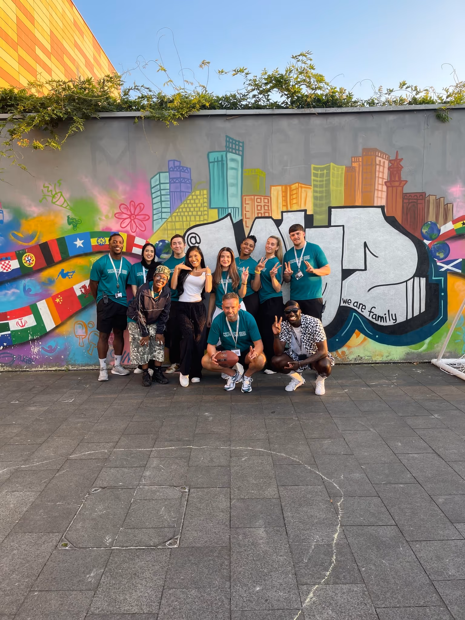 Group of diverse young people posing cheerfully in front of a colorful graffiti wall with the text '@mcr we are family'.