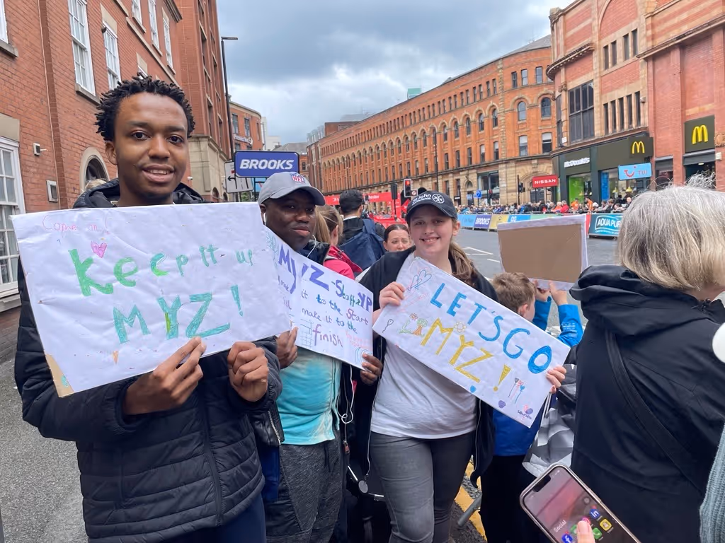 Three people holding handmade signs with supportive messages for MYZ at a street event with a crowd and brick buildings in the background.