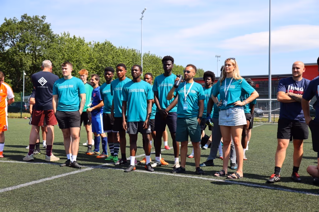 Group of people standing on a soccer field, with several wearing matching teal shirts, including one person speaking into a microphone.