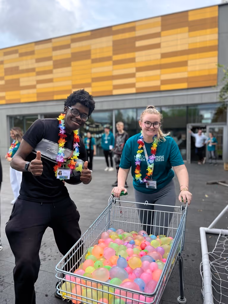 Two smiling volunteers, one giving thumbs up and the other pushing a shopping cart filled with colorful water balloons outdoors.