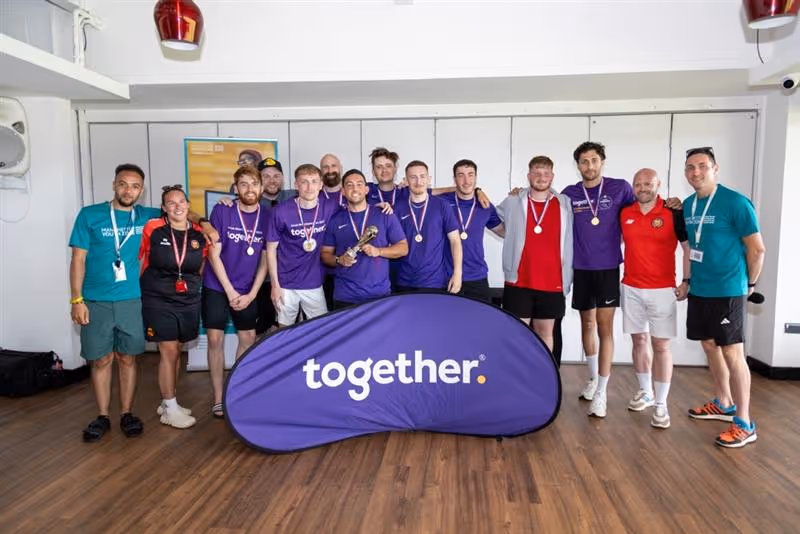 Group of smiling people standing indoors behind a purple banner with the word 'together', some wearing medals.