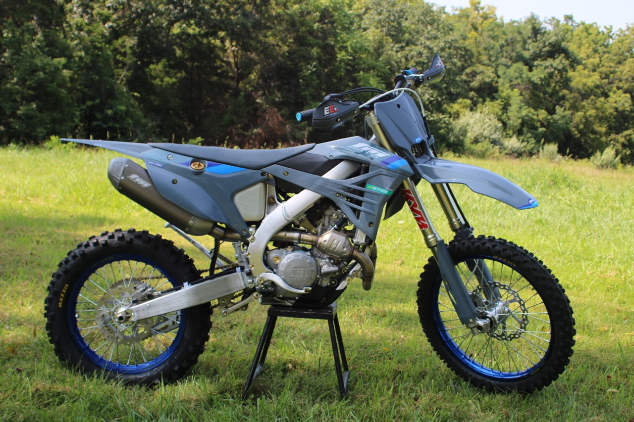 Gray dirt bike with blue rims on a grass field with trees in the background.