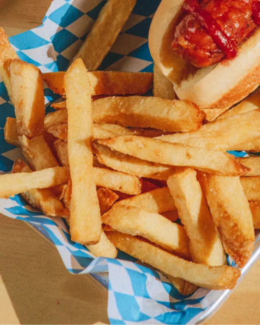 Golden French fries served in a blue and white checkered paper-lined basket with a hot dog topped with ketchup.