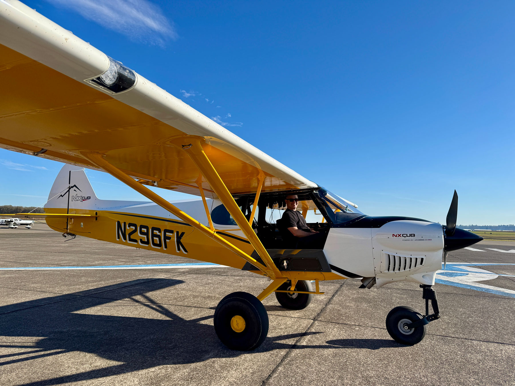 Yellow and white NX Cub airplane with pilot sitting inside on an airport tarmac under blue sky.