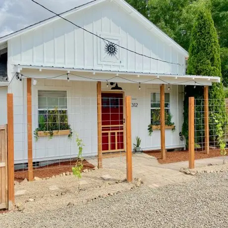 White cottage with a red door, two windows with wooden planters, a sun decor above door, and trellis fence.
