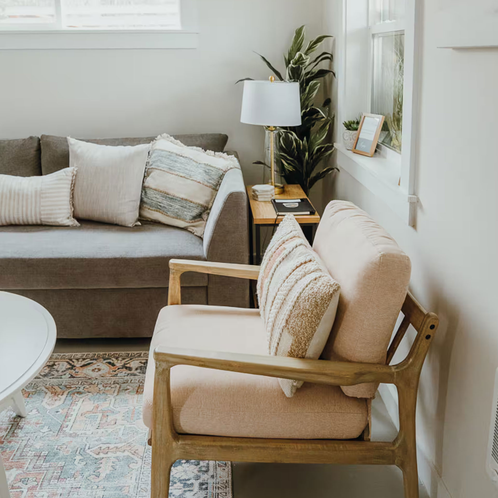 Cozy living room with beige armchair, gray sofa with cushions, wooden side table, lamp, and plant.