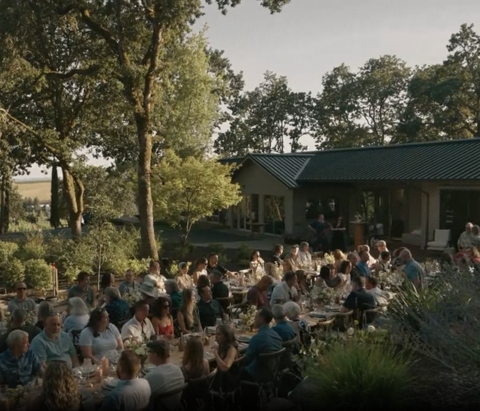 Large group seated outdoors for a meal at long tables surrounded by trees and greenery.
