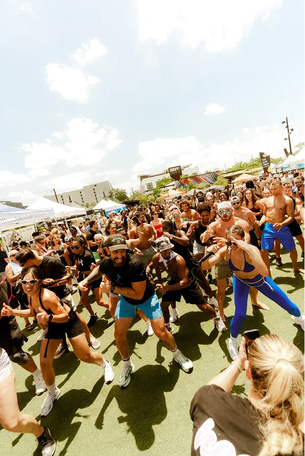 Energetic group dancing outdoors on sunny day at a lively event with tents and crowd in background.