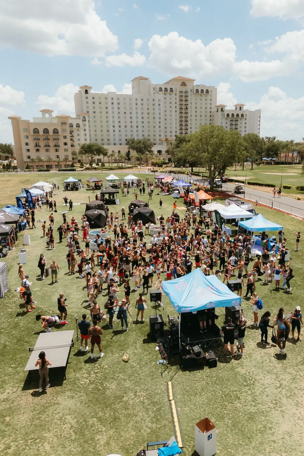 Crowd at outdoor event with tents and a stage on grass under a partly cloudy sky by a large hotel.
