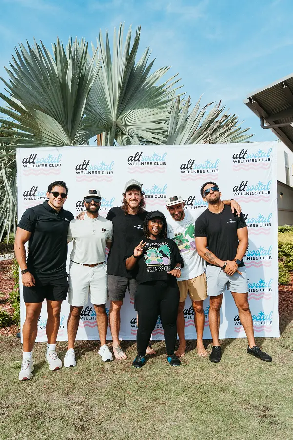 Six smiling adults posing outdoors on grass in front of a backdrop with 'alt social wellness club' logos, with large palm leaves visible behind the backdrop.