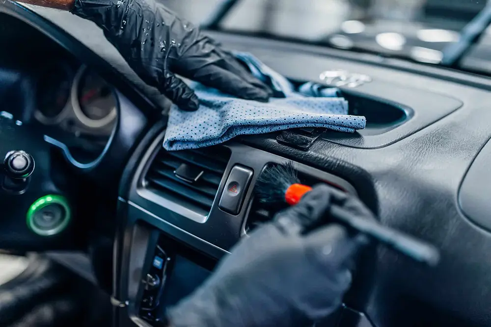 Person wearing black gloves cleaning a car dashboard with a blue microfiber cloth and a small brush.