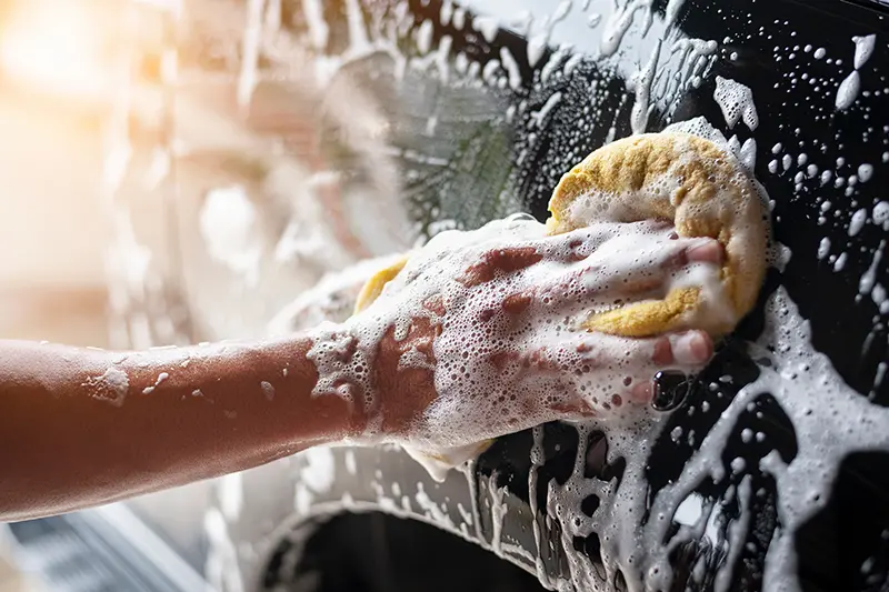 Hand covered in soap suds cleaning a black car surface with a yellow sponge.