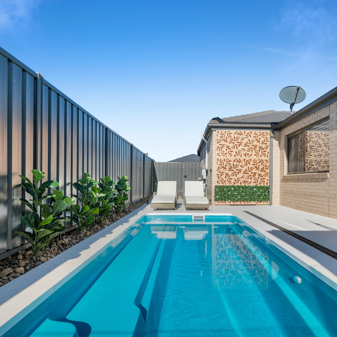 Small rectangular blue pool with two loungers beside a house and metal fence with plants.