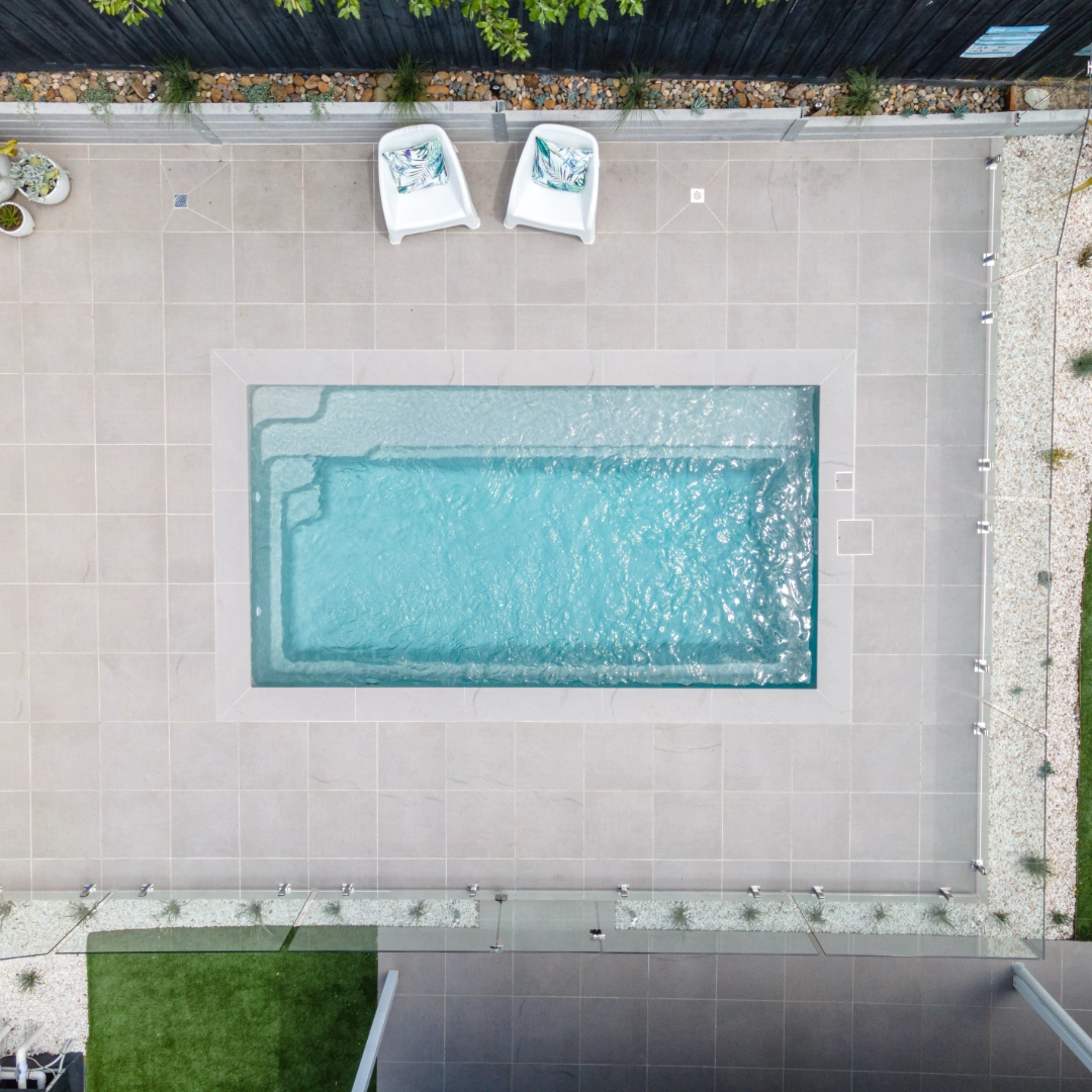 Top-down view of a rectangular pool with light tiles, two white chairs, and surrounding glass fence.