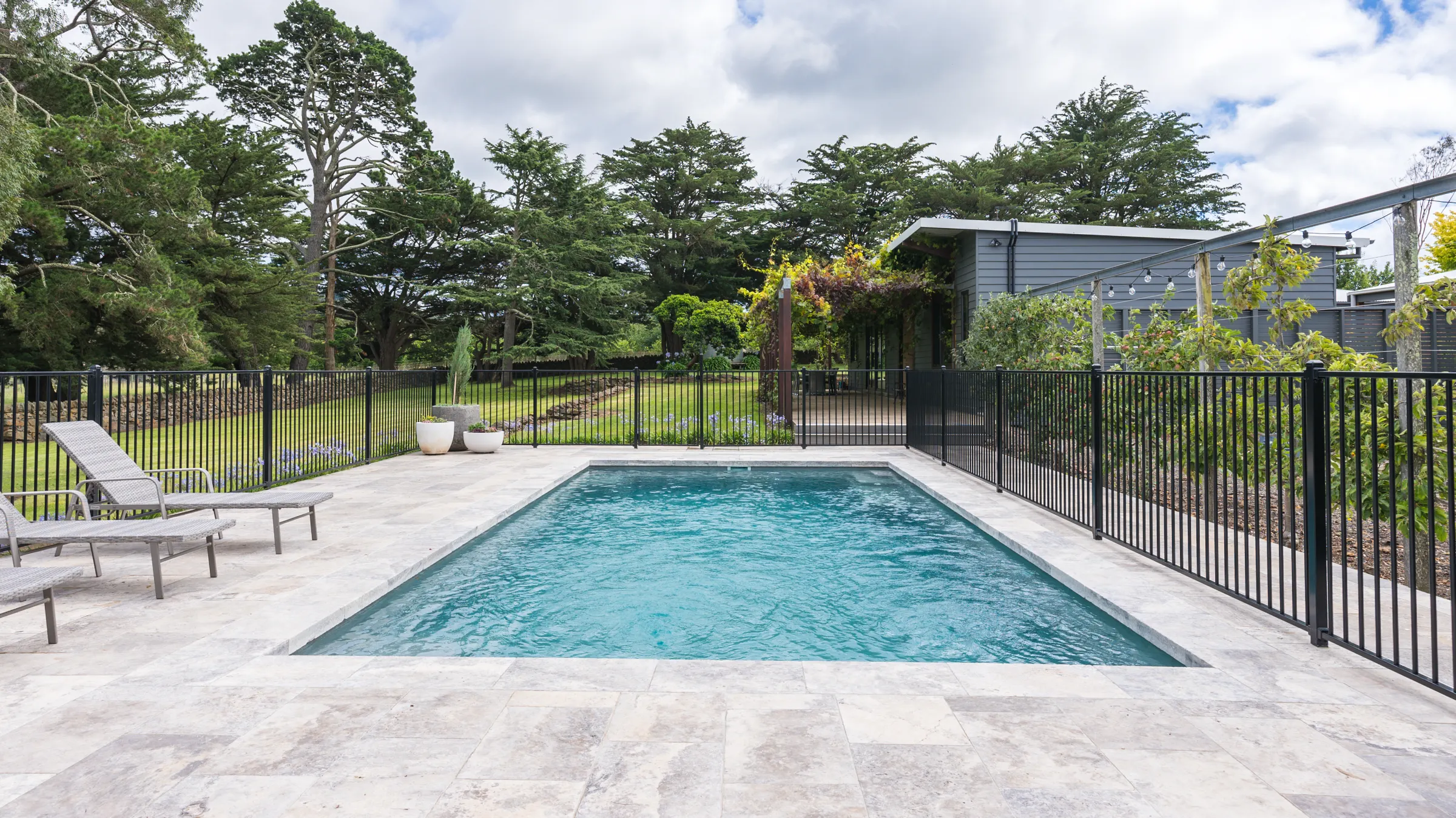 Modern rectangular pool with light stone deck and two lounge chairs in a lush green yard.
