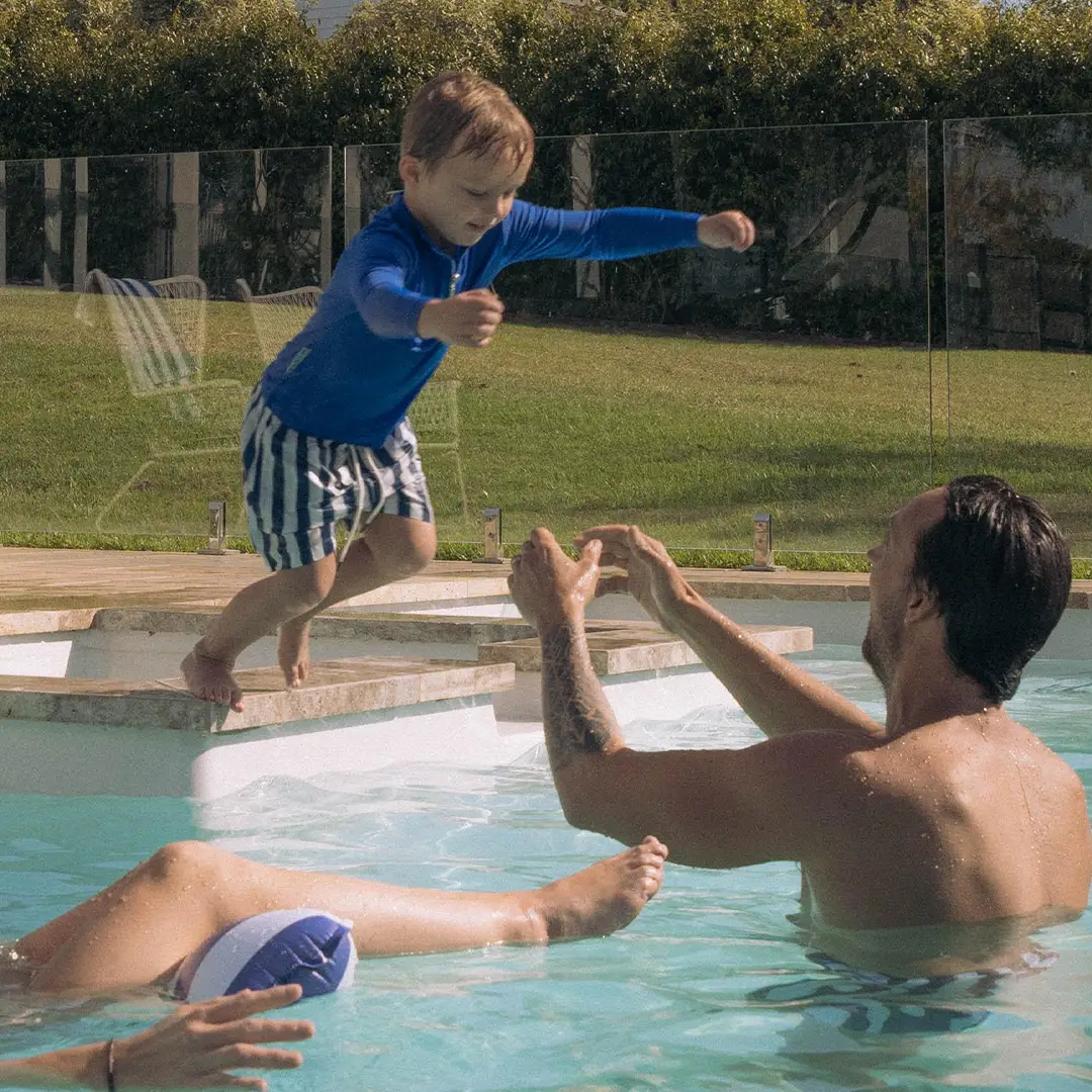 Young boy in blue shirt jumping into pool towards a man with outstretched arms ready to catch him.