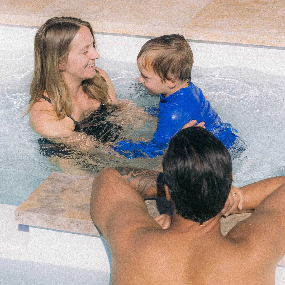 Woman in pool smiling at a young boy in a blue swim shirt with a man leaning on the pool edge nearby.