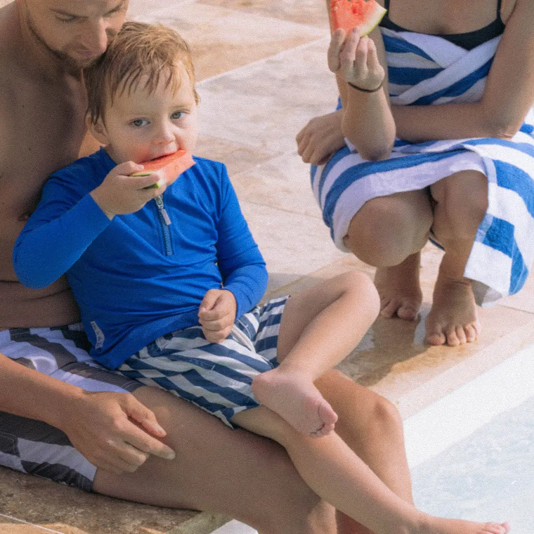 Child in blue shirt eating watermelon sitting on man's lap by pool with woman nearby in towel.