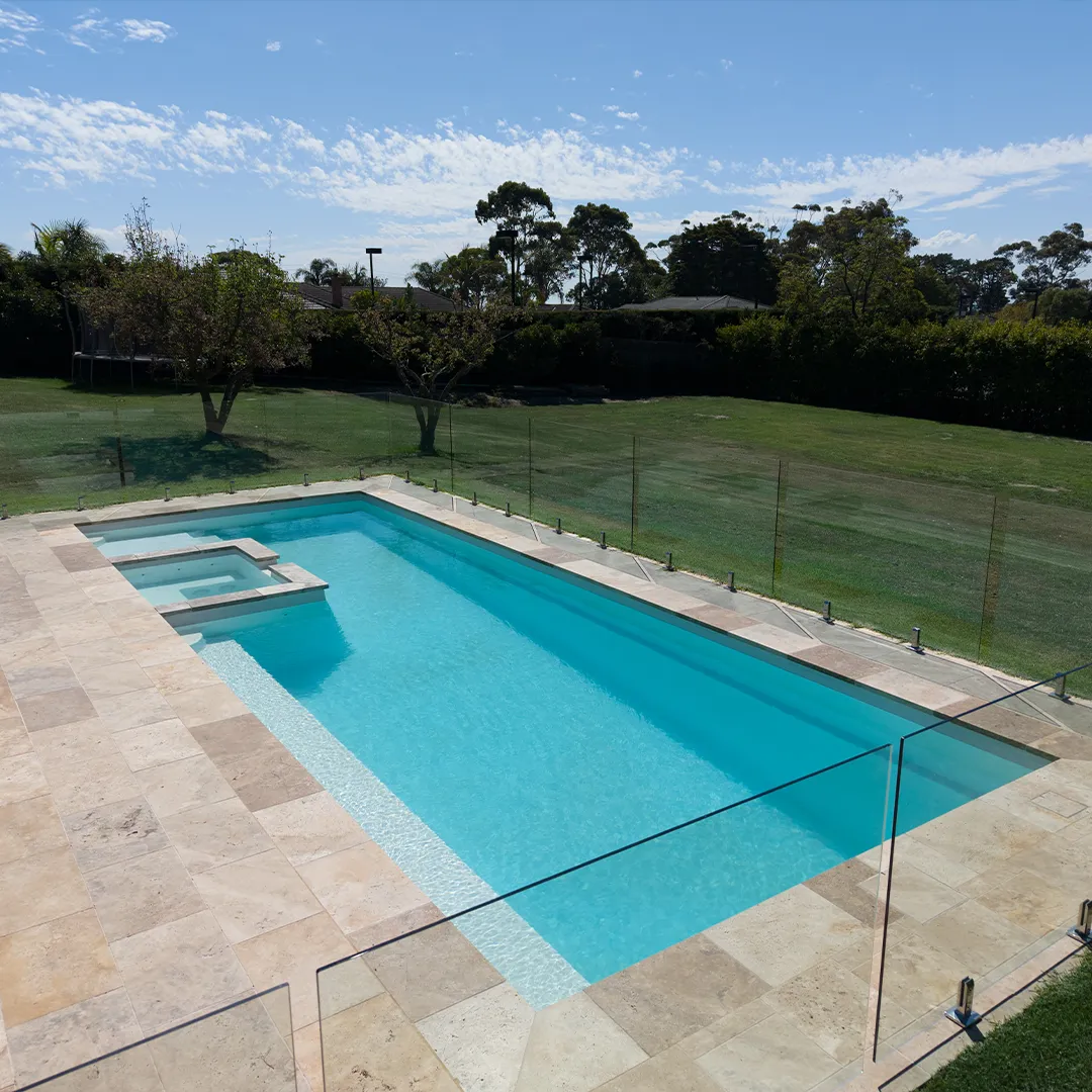Peninsula pool with attached spa, surrounded by tan stone tiles and glass fencing.
