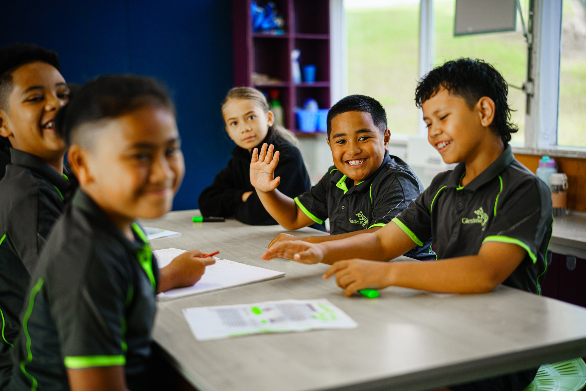Primary schools students sit at their desks, smiling and waving.