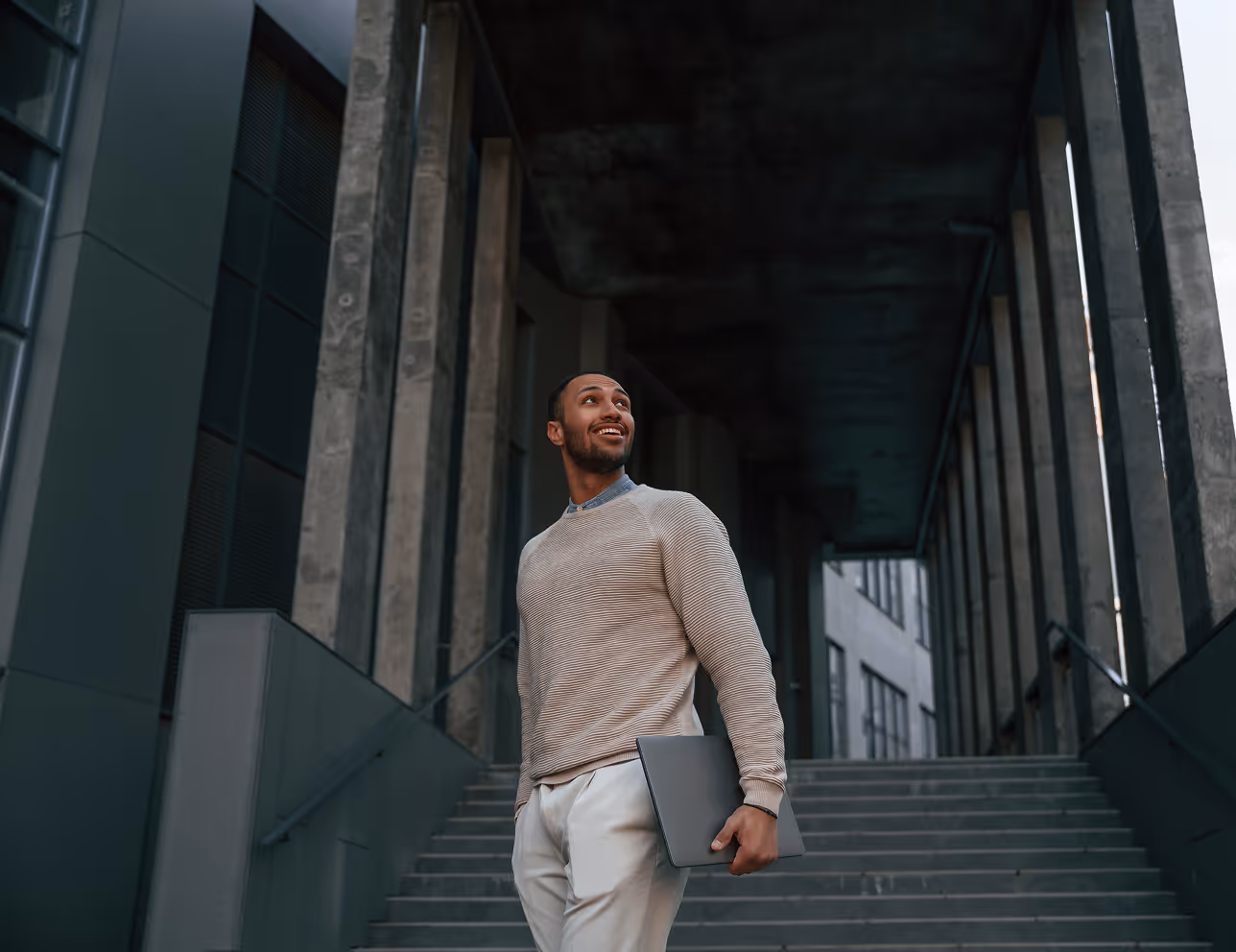 Jeune homme souriant tenant un ordinateur portable debout devant des escaliers dans un bâtiment moderne.