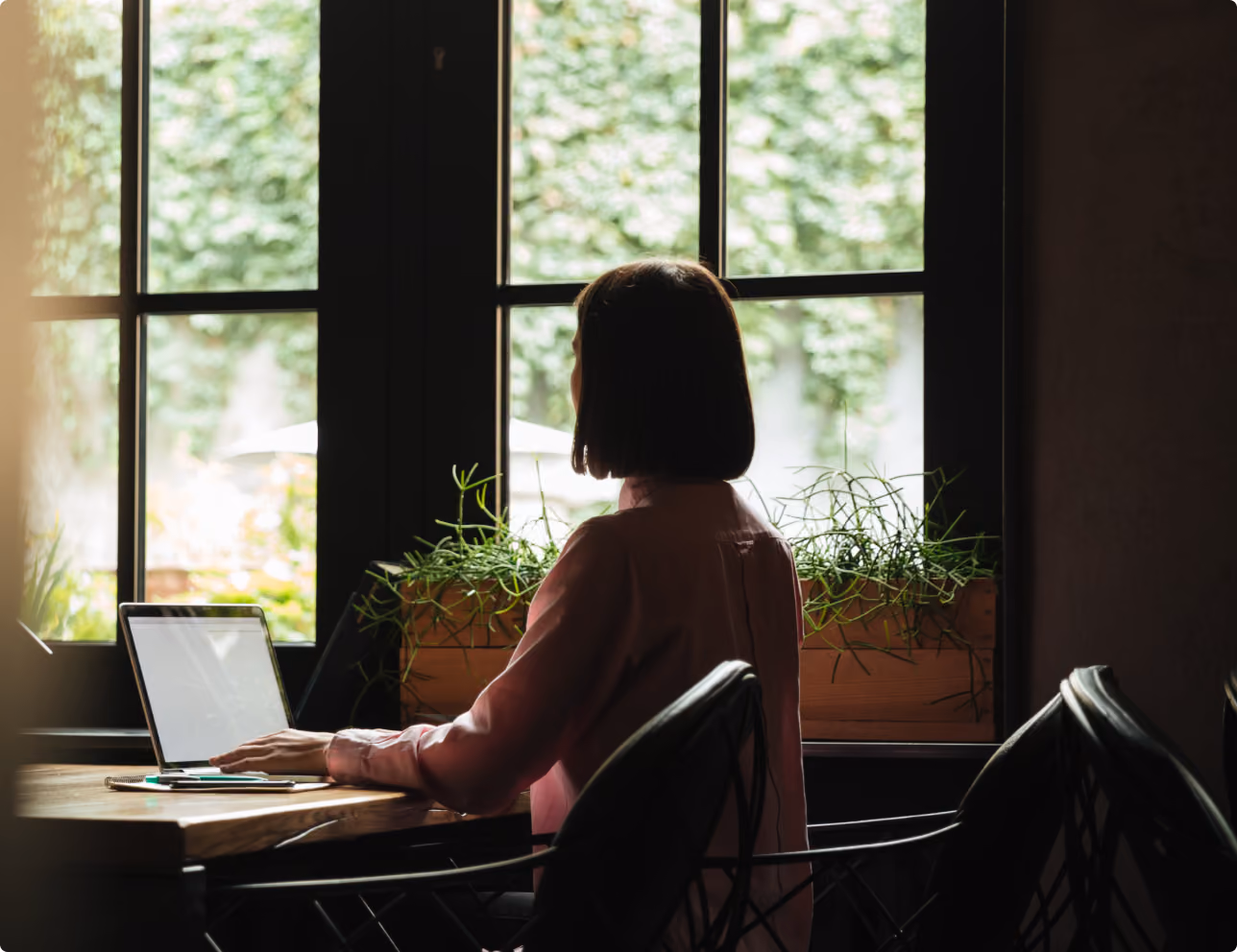 Person assise à un bureau près d'une fenêtre avec un ordinateur portable et des plantes vertes.