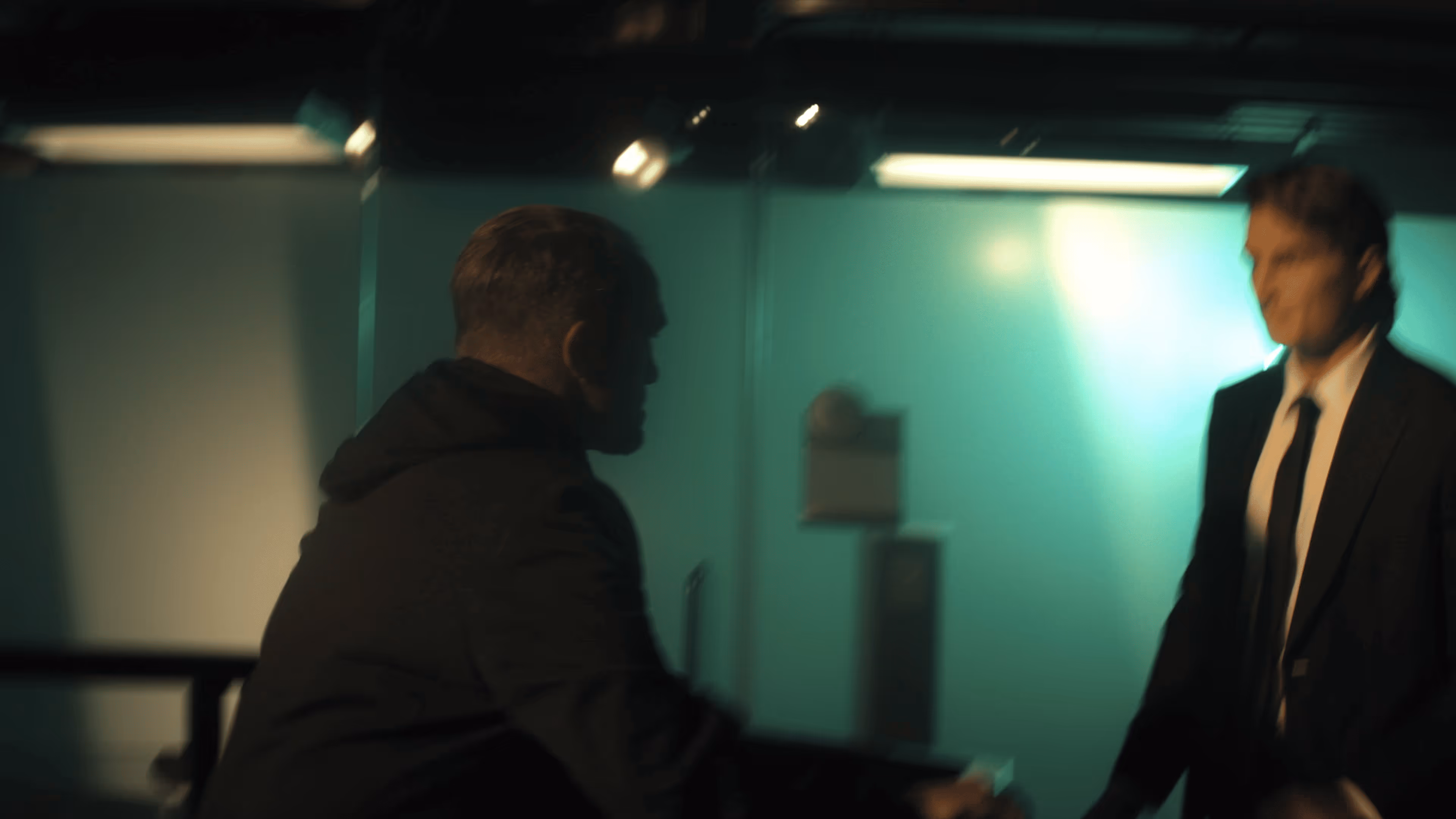 Two men facing each other in a dimly lit room with teal walls, one in a suit and the other in a dark jacket.
