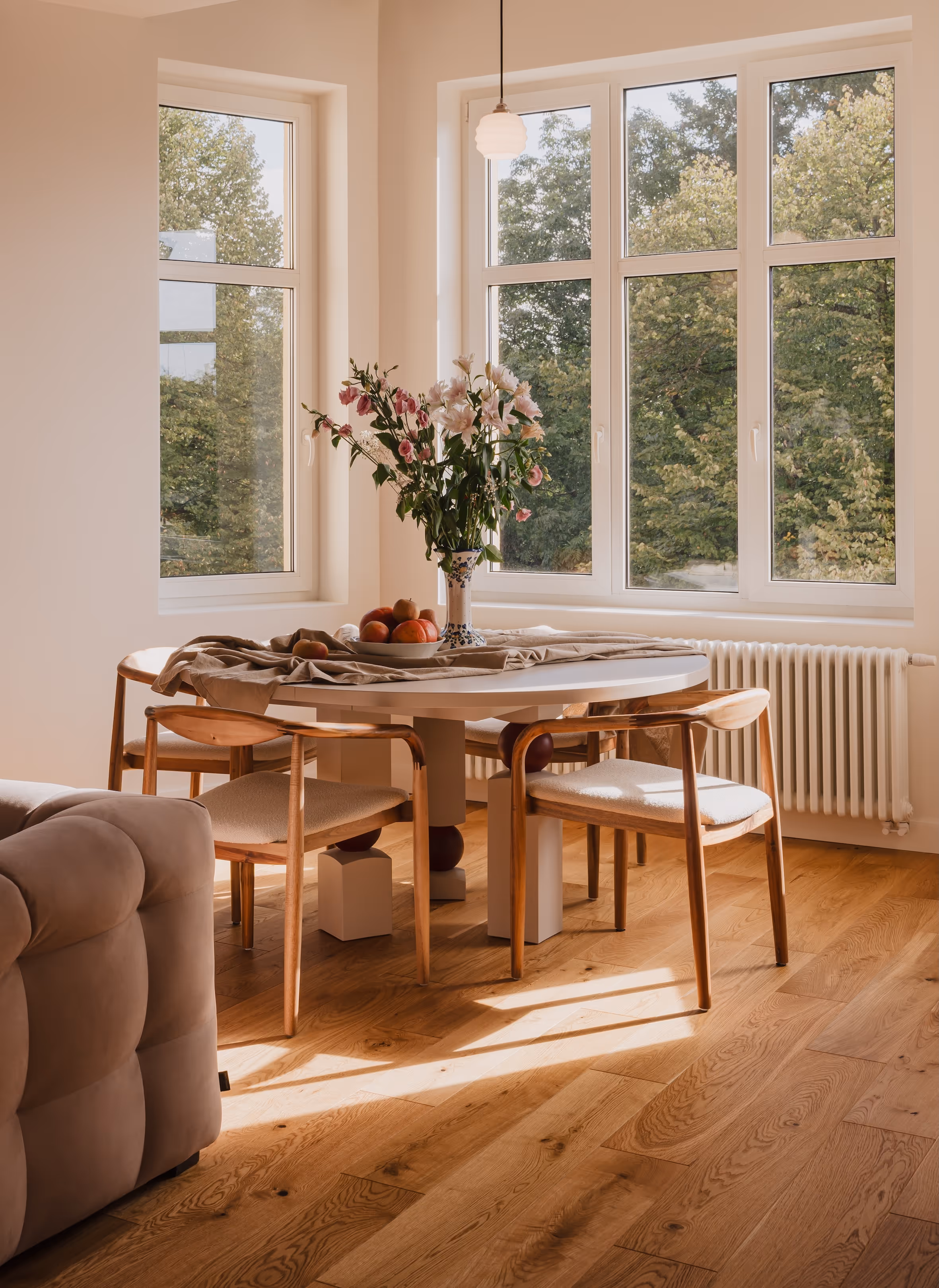 Jadalnia z okrągłym drewnianym stołem robionym na zamówienie ustawionym przy dużych oknach, z bukietem kwiatów i owocami na stole, drewniana podłoga i krzesła.