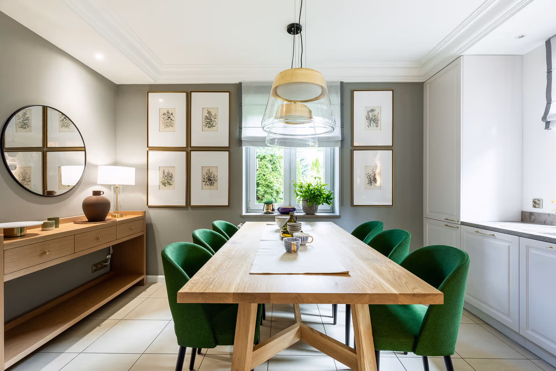 Jasna jadalnia z drewnianym stołem i zielonymi krzesłami, ozdobiona grafikami roślin na ścianie i nowoczesnym oświetleniem.