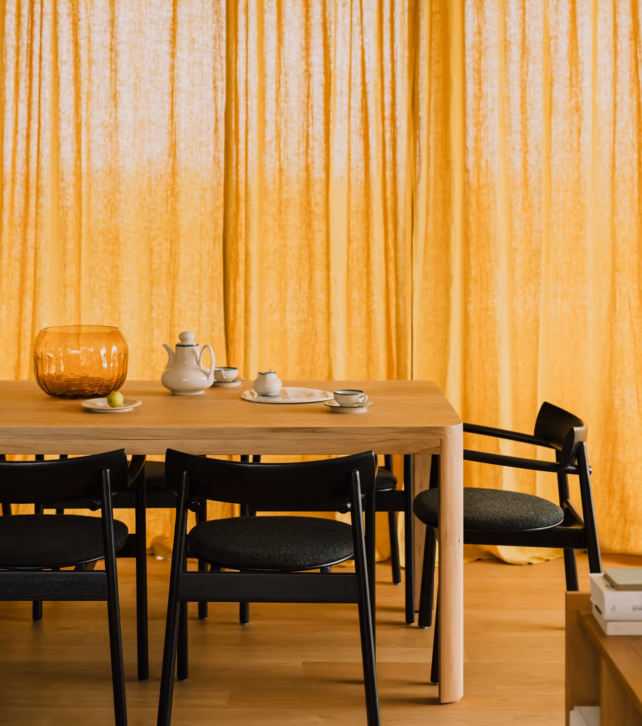 Jasny drewniany dębowy stół rozkładany z czarnymi krzesłami na tle żółtych zasłon, na stole porcelanowy zestaw do herbaty i pomarańczowa przezroczysta misa.