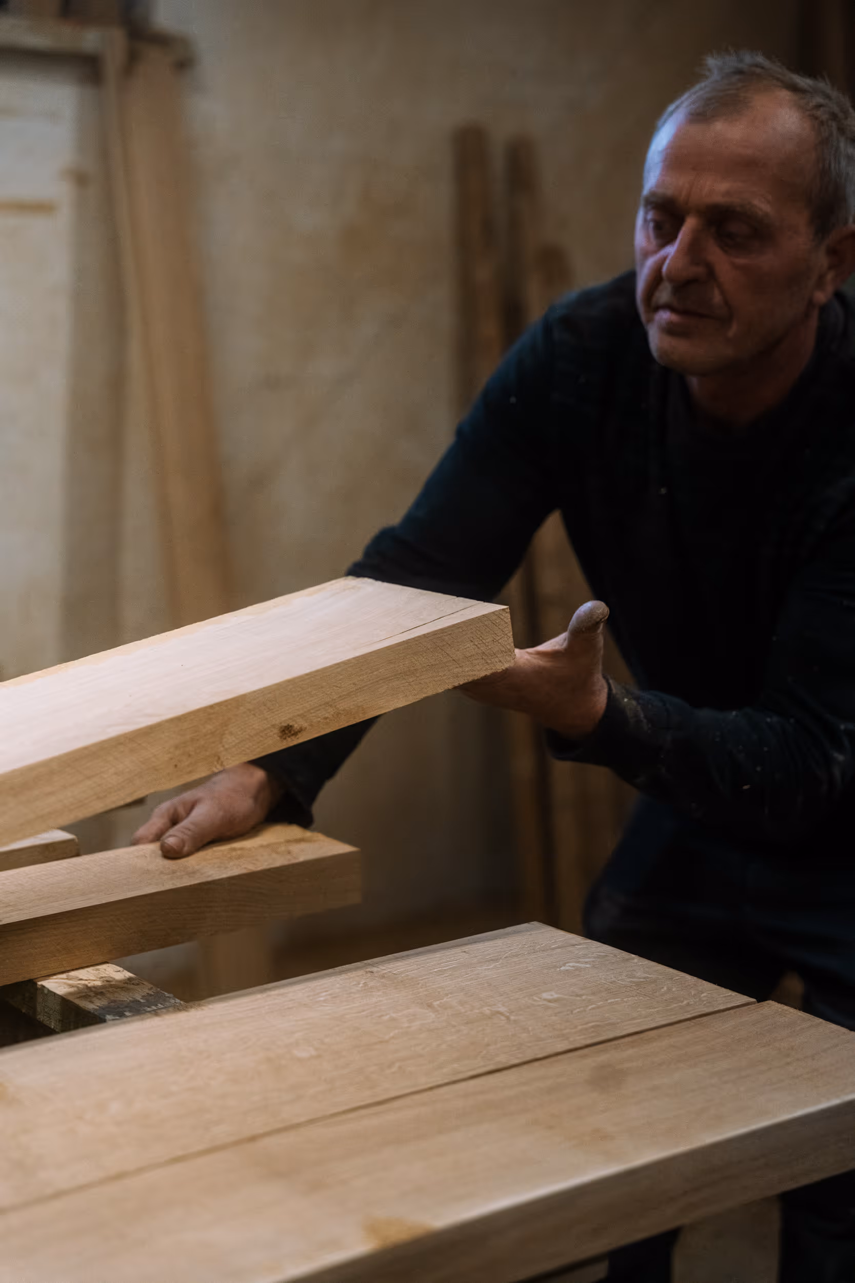 Mężczyzna w warsztacie stolarskim trzyma deskę z litego drewna, obok leżą kolejne deski na stole roboczym.